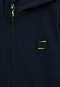 Marineblaue Kapuzenjacke mit Reißverschluss, verziert mit grünen Kordakzenten und einem rechteckigen Logopatch mit gelbem Text auf der Vorderseite.