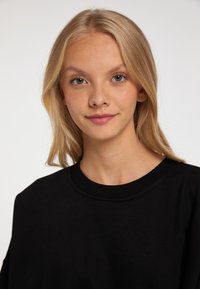 Felpa nera con scollo rotondo e polsini a costine, caratterizzata da una texture morbida. I capelli del modello sono lunghi e lisci, incorniciando il viso.