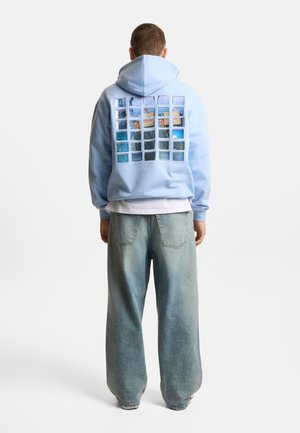 Mężczyzna noszący jasnoniebieską bluzę z kapturem z siatką małych zdjęć nadrukowanych na plecach, w połączeniu z luźnymi, jasnymi dżinsami.