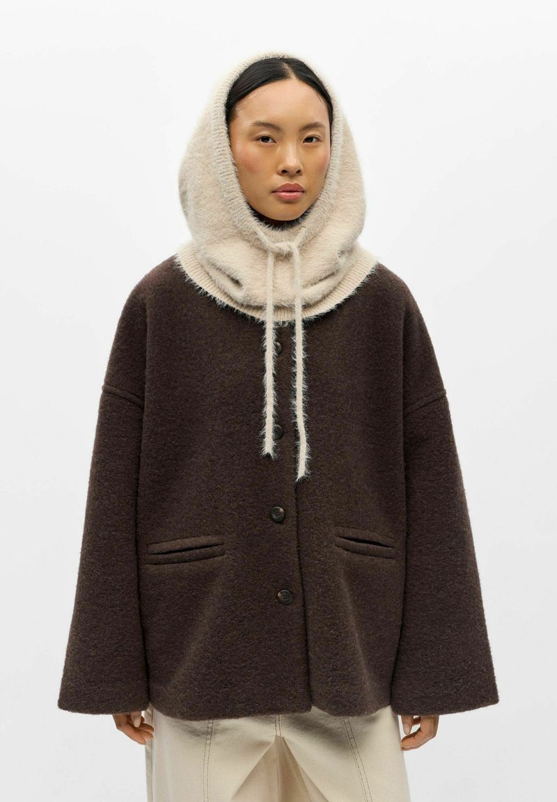 Femme portant un manteau marron foncé boutonné et une capuche beige clair duveteuse avec des cordons sur la tête.