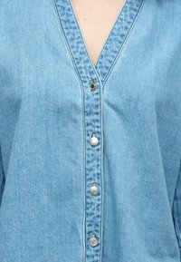 Ανοιχτό μπλε τζιν πουκάμισο με V-λαιμόκοψη και τέσσερα ορατά ασημένια κουμπιά snap κατά μήκος του κέντρου μπροστά.