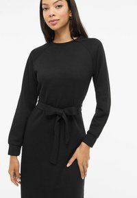 Černé žebrované pletené šaty s dlouhými rukávy, kulatým výstřihem a páskem na zavazování v pase. Mají volný střih a texturovanou látku.