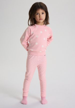 Jong meisje staat met handen achter de rug, draagt een roze sweatshirt met witte stippen en bijpassende legging, met roze sokken, tegen een effen achtergrond.