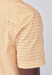 Camicia a maniche corte di colore arancione con un motivo a griglia bianca, realizzata in tessuto morbido, caratterizzata da un orlo arrotondato e dettagli di cucitura sottili.
