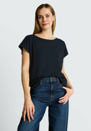 Young woman with straight blonde hair wearing a black short-sleeve top tucked into blue high-waisted jeans, standing against a plain background.