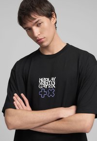 T-shirt nera in cotone con testo sovrapposto in bianco e blu, accompagnato da simboli grafici "+" e "X". Vestibilità comoda con scollatura a girocollo.