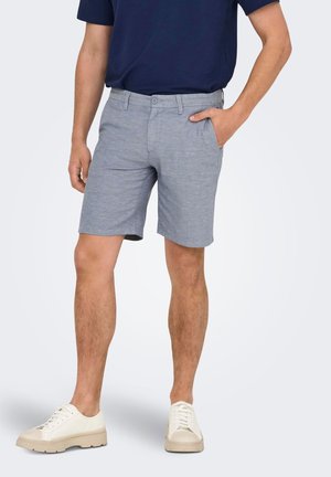 Homme portant un short gris clair à boutons, chemise bleu marine, baskets blanches, une main dans la poche, debout devant un fond uni.