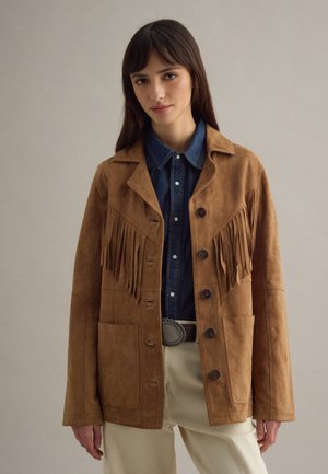 Frau trägt eine braune Wildlederjacke mit Fransen über einem blauen Jeanshemd und beigen Hosen mit dekorativer Gürtelschnalle, stehend vor einem schlichten Hintergrund.