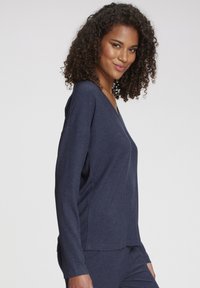 Woman with curly hair wearing a navy blue ribbed long-sleeve top and matching pants, smiling slightly, against a plain white background.
