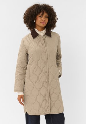 Femme aux cheveux bouclés portant un manteau beige matelassé avec un col sombre et un haut à col roulé blanc, debout devant un fond uni.