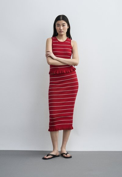 Γυναίκα που στέκεται με σταυρωμένα τα χέρια, φορώντας αμάνικο κόκκινο midi φόρεμα με λευκές ρίγες και μαύρα σανδάλια, μπροστά σε απλό γκρίζο φόντο.