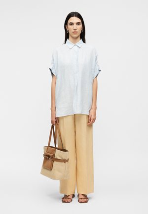 Sieviete ar gariem tumšiem matiem nēsā gaiši zilu īsroku svītrainu kreklu, bēša krāsas plašus bikses, brūnas sandales un nes bēša krāsas iepirkumu maisiņu ar brūnām siksnām.