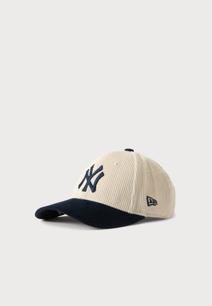 Kordfløjls baseballkasket med cremefarvet krone og sort skygge med mørkt logo broderi og lille sideemblem på hvid baggrund.