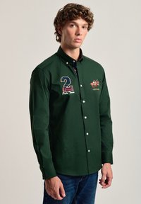 Camisa de manga larga verde, con botones, que presenta el número bordado "2", el texto "Polo Team" y un gráfico de un caballo en el pecho, hecha de una tela suave.