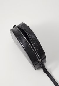 Bolso circular negro de piel sintética con textura de cocodrilo, cuenta con un cierre de cremallera plateado y una correa ajustable.