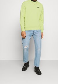 Man med ljusgrön Nike-tröja, ljusblå slitna jeans, svarta sneakers och en guldhalskedja mot en enkel vit bakgrund.