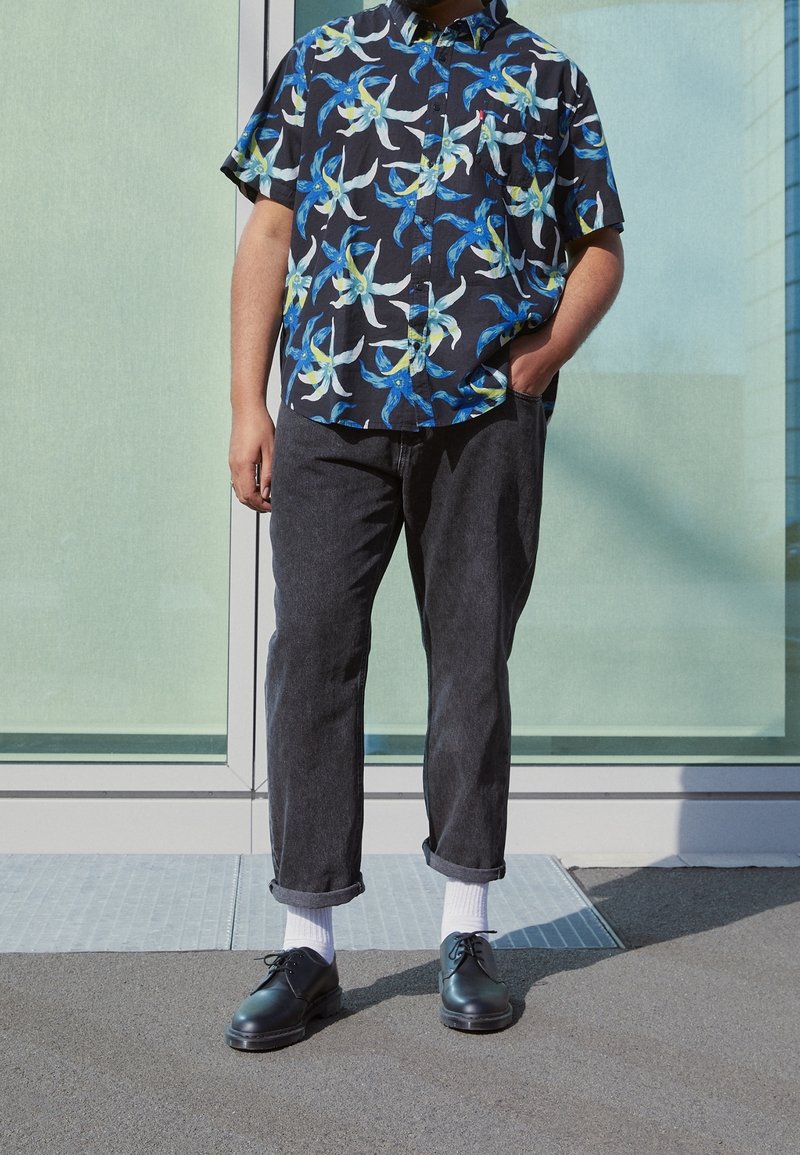 Chemise à manches courtes avec motif floral bleu et blanc, associée à un pantalon gris foncé retroussé et des chaussures noires, portée avec des chaussettes blanches.