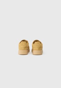 Zapatillas de ante amarillas con un perfil bajo, que cuentan con un acabado texturizado y una marca grabada en el talón, además de detalles de diseño sutiles.