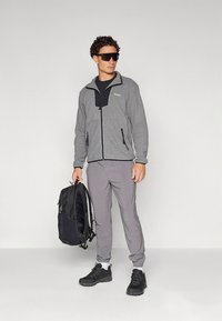 Pilko spalvos flisinė striukė su juodais akcentais, kišenės su užtrauktukais; pilkos kelnės; juodi sportbačiai; modelis nešioja juodą kuprinę ir dėvi akinius nuo saulės.