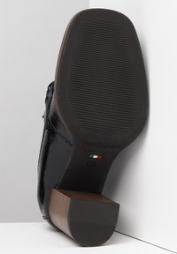 Zapato de charol negro con suela de goma texturizada y tacón cuadrado marrón. Presenta detalles de marca y la bandera italiana en la suela.