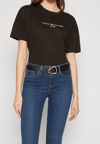 Mujer vestida con una camiseta negra de Tommy Hilfiger metida en unos jeans azules, con un cinturón negro que tiene una hebilla triangular dorada, de pie frente a un fondo blanco.
