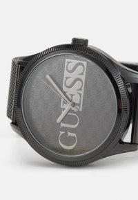 Black mesh band watch with a round case, a transparent watch face featuring a subtle gray patterned background, and prominent "GUESS" branding.