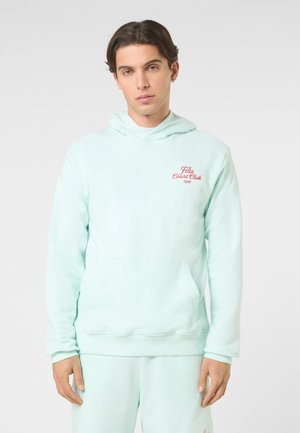 Giovane uomo con una felpa con cappuccio azzurro chiaro con la scritta "Fila Court Club 1911" in rosso, in piedi contro uno sfondo chiaro e uniforme.