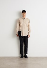 Beige Langarmhemd mit Knöpfen, kombiniert mit schwarzen Hosen und Lederschuhen. Hält eine kleine schwarze Clutch. Minimalistisches Design.