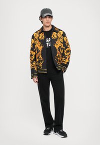 Bărbat îmbrăcat cu o jachetă neagră cu model auriu, pantaloni negri, cămașă neagră cu text alb, adidași negri și albi și o șapcă gri închis.