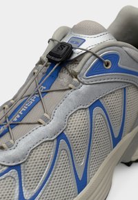 Αθλητικό παπούτσι με γκρι επάνω μέρος από διχτυωτό υλικό, μπλε λεπτομέρειες και γκρι υφή ενσωματωμένη ενδιάμεση σόλα από καουτσούκ. Διαθέτει σύστημα γρήγορου κορδονιού για εύκολη προσαρμογή.
