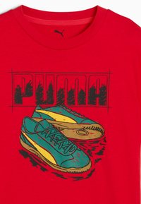 Rode T-shirt met een grafische afbeelding van turquoise en gele schoenen, met opvallende "PUMA" tekst erboven in zwart, die het ontwerp en de levendige kleuren benadrukt.