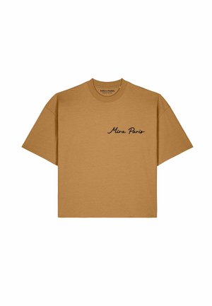 Frau trägt beiges T-Shirt mit schwarzer abstrakter Gesichts-Linienzeichnung auf dem Rücken, blaue Jeans und Schildpatt-Sonnenbrille und steht auf einer Kopfsteinpflaster-Straße.
