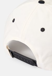 Krämfärgad baseballkeps med en texturerad tyg, svarta sömddetaljer och ett justerbart svart plastband bak.