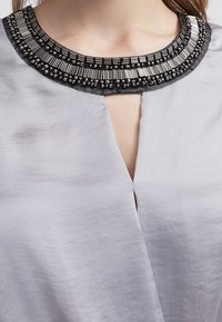 Blouse gris clair avec un décolleté profond, ornée d'un col perlé noir et argenté. Texture lisse, design élégant, et détails complexes en perles.