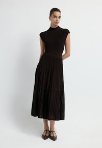 Žebrované černé šaty s krátkými rukávy, dobře padnoucím trupem a volnou plisovanou sukní. Mají lesklou texturu a končí zhruba v polovině lýtek.