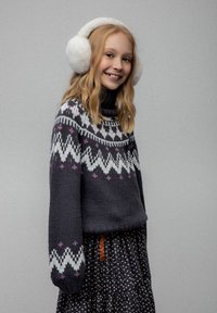 Uśmiechnięta dziewczyna z długimi blond włosami, nosząca białe puszyste nauszniki, ciemny sweter z białymi i fioletowymi wzorami oraz kwiatową spódnicę.