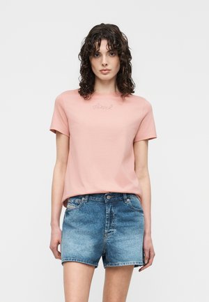 Frau mit lockigem dunklem Haar trägt ein hellrosa Kurzarmoberteil und blaue Jeansshorts, steht vor einem schlichten hellgrauen Hintergrund.