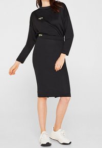 Femme portant un haut à manches longues noir et une jupe assortie jusqu'aux genoux avec des baskets blanches, se tenant devant un fond uni.