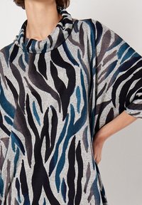 Pull gris à col châle avec un design fluide, présentant des rayures zébrées noires, bleu foncé et bleu clair. Tissu doux avec une coupe décontractée.