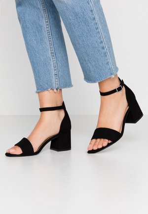 Sandales à talons hauts en suède noir avec un talon carré épais, bout ouvert et bride de cheville, présentées avec un jean bleu clair à ourlet effiloché.