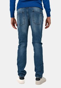 Jeans in denim blu da uomo con una vestibilità slim, dotati di due tasche posteriori e una leggera sbiaditura. Abbinati a sneakers bianche e a un maglione blu.