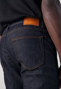 Jean en denim foncé avec coutures orange, doté d'une étiquette en cuir SANDRO à la taille arrière et de poches arrière standard.