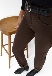 Pantaloni in cotone marroni con taglio dritto, dotati di tasche frontali e una cintura nera texturizzata con fibbia argentata. Presenti stivaletti neri alla caviglia.