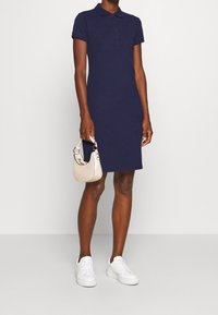 Vestido de polo azul marino con mangas cortas y tapeta de botones. Combinado con zapatillas blancas y un pequeño bolso beige. Textura de tela suave.