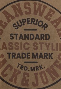 La tela marrón presenta texto audaz en negro y marrón, mostrando el nombre de la marca "JACK & JONES", con frases como "SUPERIOR", "STANDARD" y "TRADE MARK".