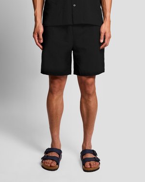 Osoba oblečená v čiernych šortkách a čiernych sandáloch s dvojitými remienkami, stojaca na jednofarebnom sivom pozadí.