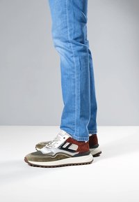 Sneakers in Olivgrün, Braun und Grau mit einem Obermaterial aus strukturiertem Mesh und Wildleder, weißer Sohle und grauen Schnürsenkeln. Kombiniert mit hellblauen Jeans.