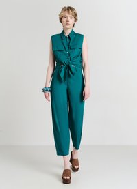 Tutina teal con design senza maniche, vita legata, due tasche frontali e gamba larga. Abbinata a sandali platform marroni e un bracciale con motivo.