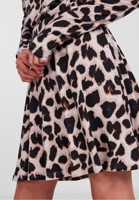 Händerna är korsade över en leopardmönstrad kjol och en matchande långärmad topp, som visar svarta och bruna fläckar på ljus beige tyg.