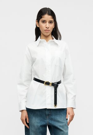 Λευκό βαμβακερό πουκάμισο με γιακά και μία τσέπη, σφιγμένο στη μέση με μια μαύρη δερμάτινη ζώνη με χρυσή αγκράφα. Μπλε τζιν παντελόνι.